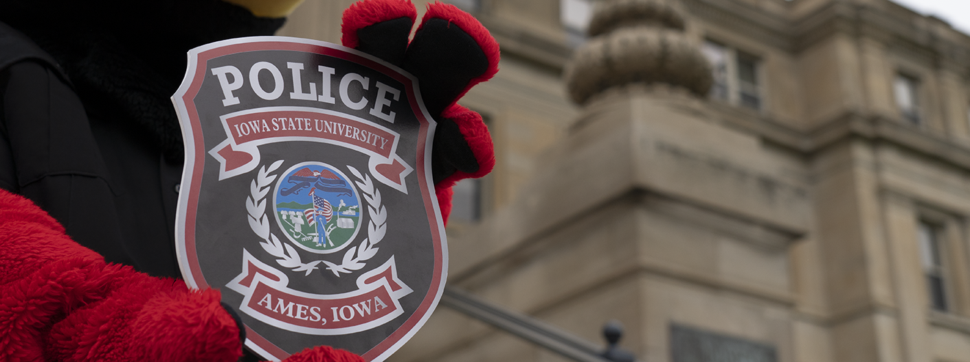 Cy mascot holds an oversized ISU Police badge outside Beardshear Hall.